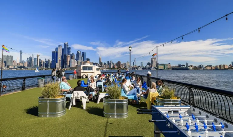 People relaxing on a riverside lawn area in Hoboken, NJ, with foosball tables, a food truck, and a stunning view of the Manhattan skyline across the Hudson River in Hoboken, NJ