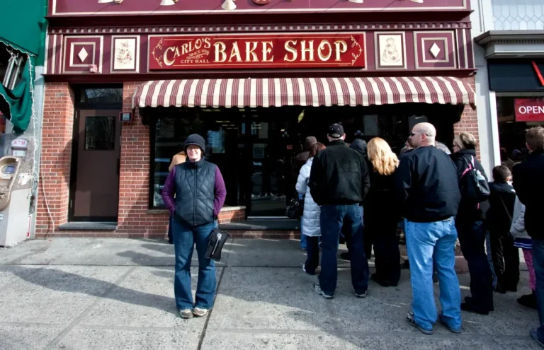 People standing in line outside the famous Carlo’s Bake Shop in Hoboken, NJ, under a red and white striped awning on a chilly day in Hoboken, NJ