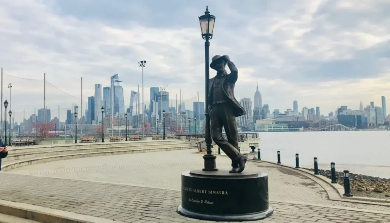 Bronze statue of Frank Sinatra leaning against a lamppost on the Hoboken waterfront, with the New York City skyline and Empire State Building in the background in Hoboken, NJ