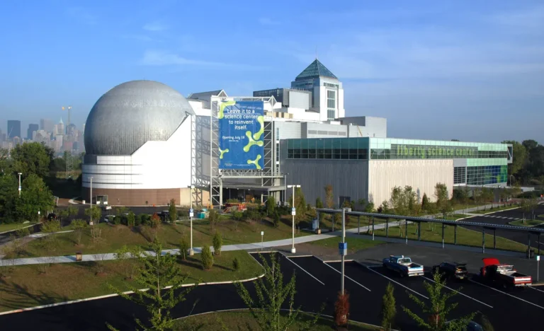 Modern science center with a large dome structure, glass-walled building, and banner display, surrounded by landscaped grounds and parking lot in Jersey City, NJ