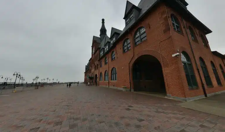 Historic red-brick building with arched windows and towers along a wide paved promenade on a cloudy day in Jersey City, NJ