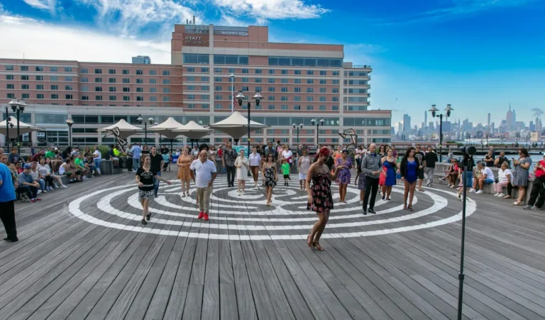 People dancing on a wooden boardwalk stage surrounded by a crowd, with a hotel and city skyline in the background on a sunny day in Jersey City, NJ