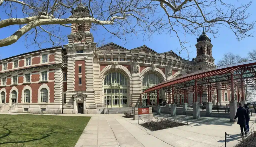 Historic red-brick and stone building with arched windows and towers, located behind a red metal canopy structure under a clear blue sky in Jersey City, NJ