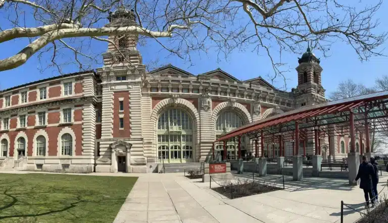 Historic red-brick and stone building with arched windows and towers, located behind a red metal canopy structure under a clear blue sky in Jersey City, NJ