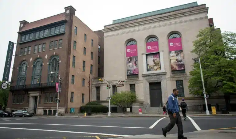 People walking across a crosswalk in front of a large museum building with banners and arched windows on a cloudy day in Kearny, NJ