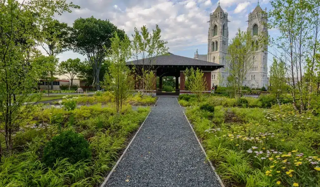 Gravel path leading through a vibrant garden to a pavilion, with a tall cathedral in the background under a partly cloudy sky in Kearny, NJ