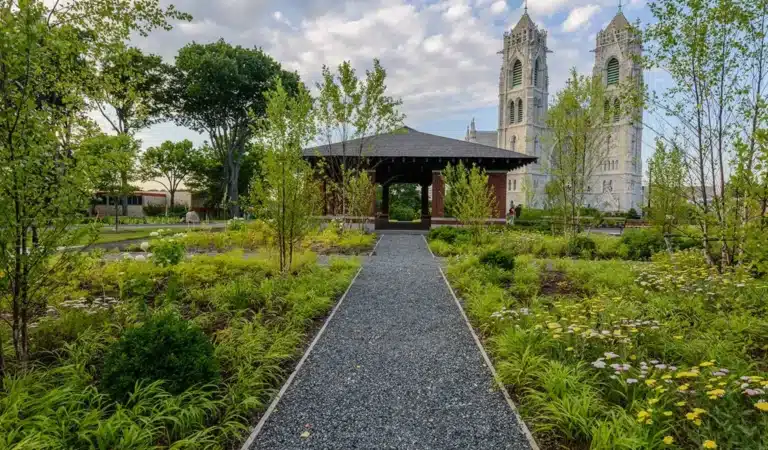 Gravel path leading through a vibrant garden to a pavilion, with a tall cathedral in the background under a partly cloudy sky in Kearny, NJ