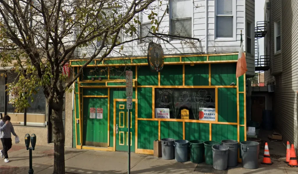 Green and yellow storefront with Irish decor, chalkboard signs in the window, trash bins out front, and a bare tree casting shadows on the sidewalk in Kearny, NJ