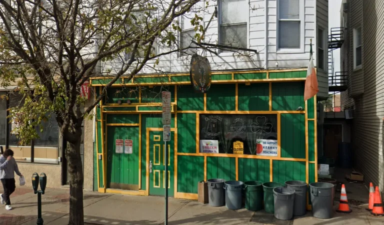 Green and yellow storefront with Irish decor, chalkboard signs in the window, trash bins out front, and a bare tree casting shadows on the sidewalk in Kearny, NJ