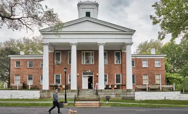 Historic brick building with tall white columns and central tower, surrounded by trees, with a person walking a small dog in front in Staten Island, NY
