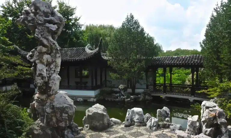 Traditional Chinese garden with ornate pavilion, stone bridge, rock formations, and lush greenery surrounding a serene pond in Staten Island, NY