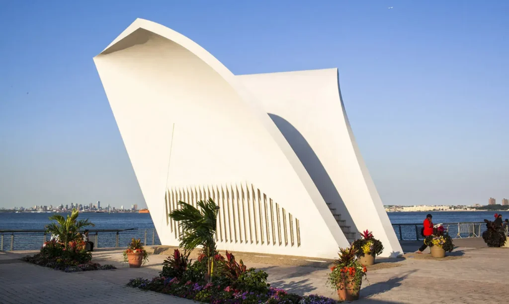 White sculptural Staten Island 9/11 Memorial with floral arrangements and waterfront view under a clear blue sky in Staten Island, NY