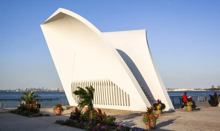 White sculptural Staten Island 9/11 Memorial with floral arrangements and waterfront view under a clear blue sky in Staten Island, NY